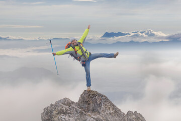 Hiker with backpack cheering elated and blissful with arms raised on top mountain foggy after...