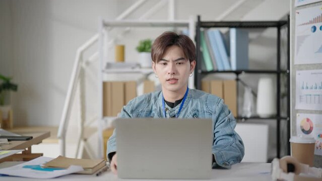 Young handsome Asian man takes a break from working on his laptop in office and gazes out of the window, lost in thought.