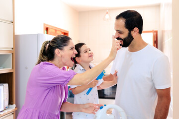 Brazilian family in the kitchen making a cake together to celebrate Father's Day. Mom playfully puts cake batter on father's nose, making everyone laugh. 