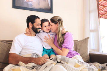 Parents and child embracing on the couch, sharing affectionate gestures. Their smiles and warmth create a joyful, heartfelt scene. Ideal for showcasing authentic, loving family interactions at home
