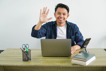 young asian freelancer sitting in workplace saluting with hand with happy expression