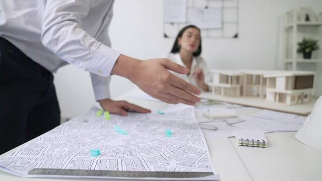 Skilled caucasian interior designer working with house model while discussing with asian architect engineer about house plan on meeting table with blueprint and house model scatter around. Immaculate.