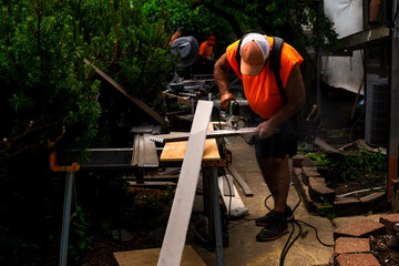 A construction worker in a bright orange shirt is cutting wood in a backyard, surrounded by tools and greenery.