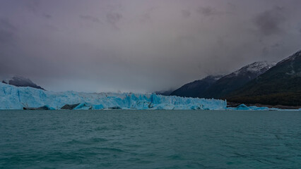 A wall of blue ice stretches across a turquoise glacial lake. Icebergs, melted ice floes float in the water. Coastal mountains in clouds and fog. Perito Moreno glacier. El Calafate. Argentina. 