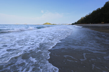 A pristine beach with turquoise waters, foamy waves, and a soft sandy shore under a blue sky with fluffy clouds.