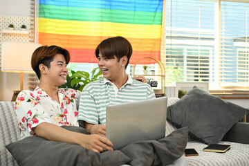 Young gay couple sitting on couch and surfing the net together at home. LGBTQ people lifestyle...