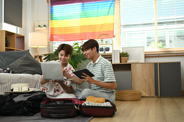 Gay couple booking hotel reservation on laptop packing suitcase on floor at home. Travel and...