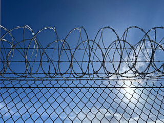 metal chain link fence with dangerous cruel sharp barbed wire against a bright blue midday sky with...