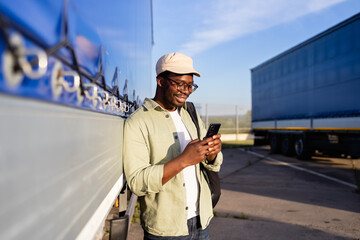 Truck driver standing by vehicle trailer and using mobile phone on parking lot.