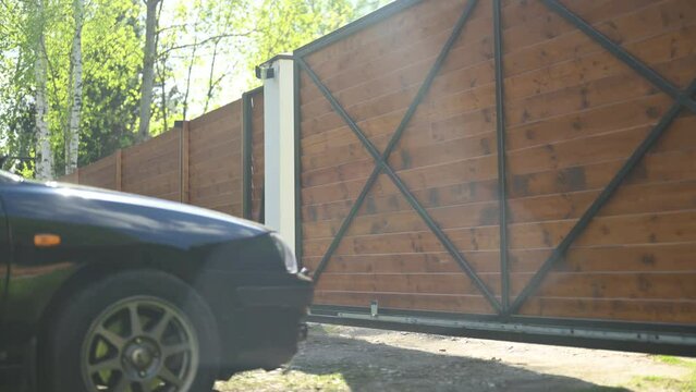 The gate of a private property opens, a black sedan drives in and out of the house on a sunny day. Long version