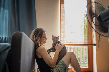 Young woman with a gray cat in her arms sitting at home on the floor in front of a fan, escapefrom...