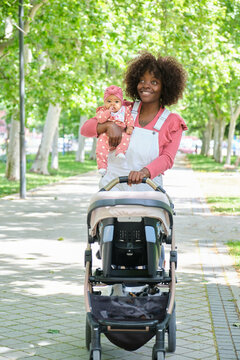 African woman walking with her baby girl pushing a stroller in a park.