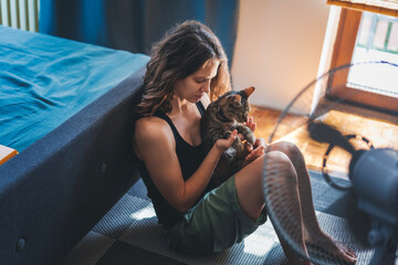 Young woman with a gray cat in her arms sitting at home on the floor in front of a fan, escapefrom the summer heat concept