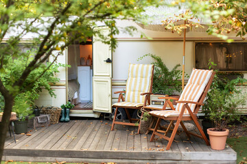 Obraz premium Cozy Campsite on caravan or camper van in forest. Trailer of mobile home stands in garden in camping. Backyard with RV house with garden furniture. Two deckchairs near outside caravan trailer. 