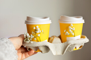 Female hand holding paper yellow cup with takeaway coffee decorated with gypsophila flower. Coffee...