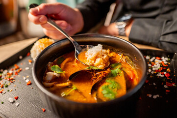 A close-up of a hand holding a spoon over a bowl of soup, garnished with herbs, with rice on the side.