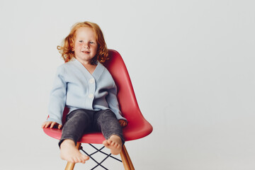 Small girl sitting on red chair against white background, portrait of a child in minimalist decor...