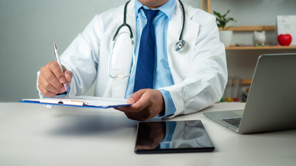 Male doctor sitting at work desk checking work plans and writing document reports in hospital office, healthcare worker and doctor service, digital laptop in modern office, medical technology concept.