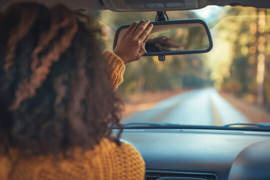 Rear view of a woman adjusting the rearview mirror in her car while driving through a scenic, sunlit autumn road