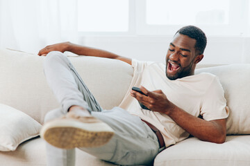 Happy young African American man sitting on a black sofa, typing a message on his mobile phone He...