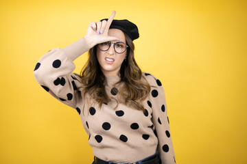 Young beautiful brunette woman wearing french beret and glasses over yellow background making fun of people with fingers on forehead doing loser gesture mocking and insulting.