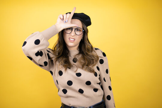 Young beautiful brunette woman wearing french beret and glasses over yellow background making fun of people with fingers on forehead doing loser gesture mocking and insulting.