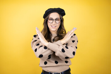 Young beautiful brunette woman wearing french beret and glasses over yellow background Rejection...