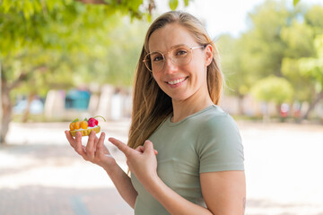 Young blonde woman holding a tartlet at outdoors and pointing it