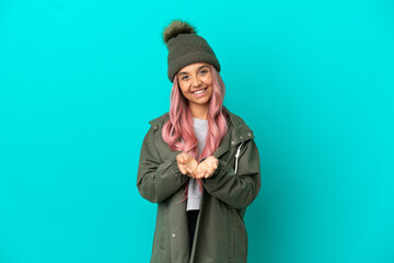 Young woman with pink hair wearing a rainproof coat isolated on blue background holding copyspace imaginary on the palm to insert an ad