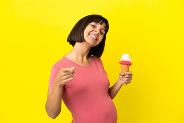 Pregnant woman holding a cornet ice cream isolated on yellow background pointing front with happy...