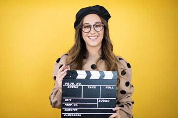 Young beautiful brunette woman wearing french beret and glasses over yellow background holding...