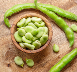 Broad beans in wooden bowl and broad bean pods near it on wooden table. Top view.