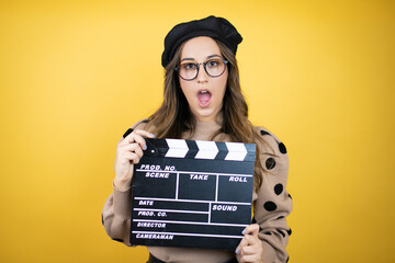 Young beautiful brunette woman wearing french beret and glasses over yellow background holding...