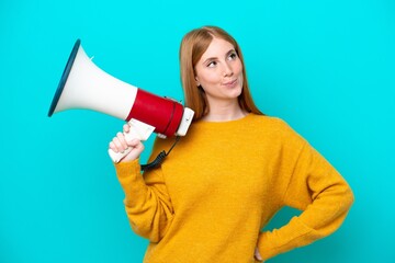 Young redhead woman isolated on blue background holding a megaphone and thinking