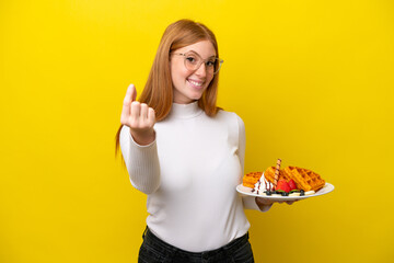 Young redhead woman holding waffles isolated on yellow background making money gesture