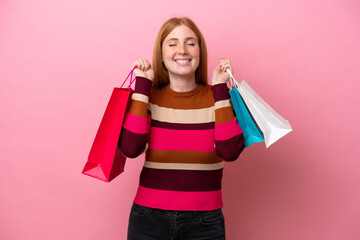 Young redhead woman isolated on pink background holding shopping bags and smiling