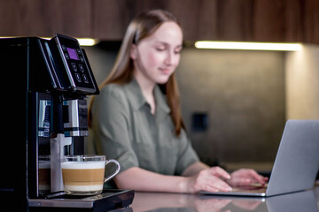 Woman drinking coffee and working on laptop near coffee machine at home.