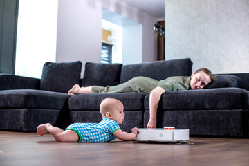 Cute young woman is sleeping on sofa in living room while little child is playing with robot vacuum...
