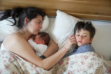Mother laying in bed with newborn baby and older son, sharing an intimate and nurturing moment. The...