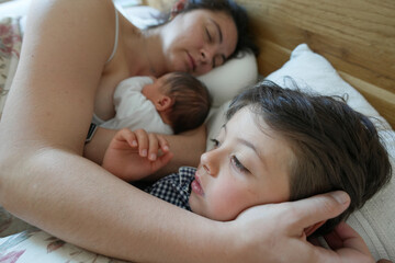 Mother cuddling newborn baby in bed, with older son close by. The serene scene captures the tender...