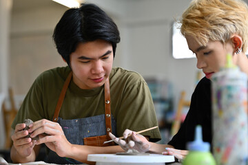 Two artists design clay pieces together with focused expressions. One artist holds a sculpting tool...