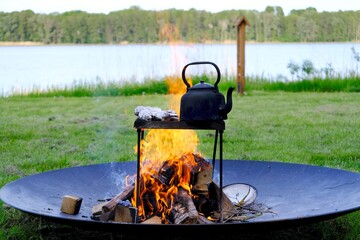 An old metal kettle stands among the flames of the fire. There's grilled food in aluminum foil....