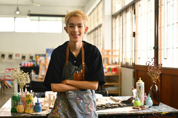 An LGBT teenage student stands confidently in a studio, smiling and ready to create. The image captures the student's enthusiasm for learning and the vibrant, supportive environment of the art space
