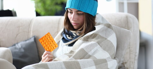 Portrait of sullen ill woman sitting indoors and wearing warm clothes to cure annoying disease. Poor girl holding yellow pack of pills and looking gloomy. Medications concept