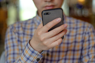 Close-up of a young East Asian man’s light-skinned hand holding a mobile phone, with his face...