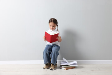 Cute little girl reading on stack of books near light grey wall