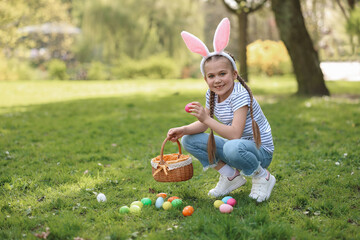 Easter celebration. Cute little girl in bunny ears hunting eggs outdoors, space for text