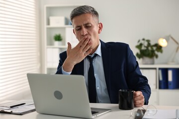 Sleepy man with cup of drink yawning at table in office