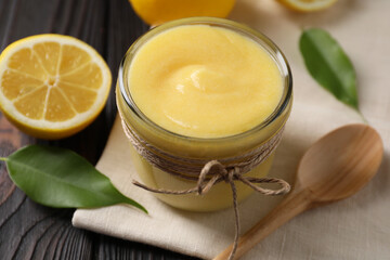 Delicious lemon curd in bowl, fresh citrus fruit, spoon and green leaves on wooden table, closeup