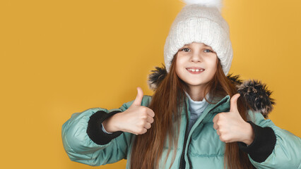 A little cheerful girl in a winter warm hat and down jacket shows a thumbs up.